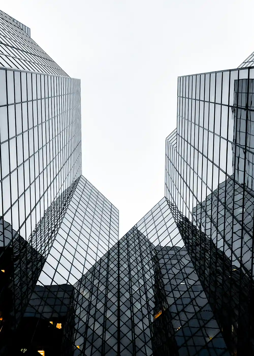 Looking up at two glass-curtain-wall skyscrapers converging symmetrically against an overcast sky